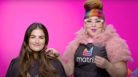 Two individuals in a salon setting with a bright pink background. One is a Matrix stylist who has colorful bangs and is wearing a pink top with ruffled sleeves, while the other is a hair model with long brown hair.